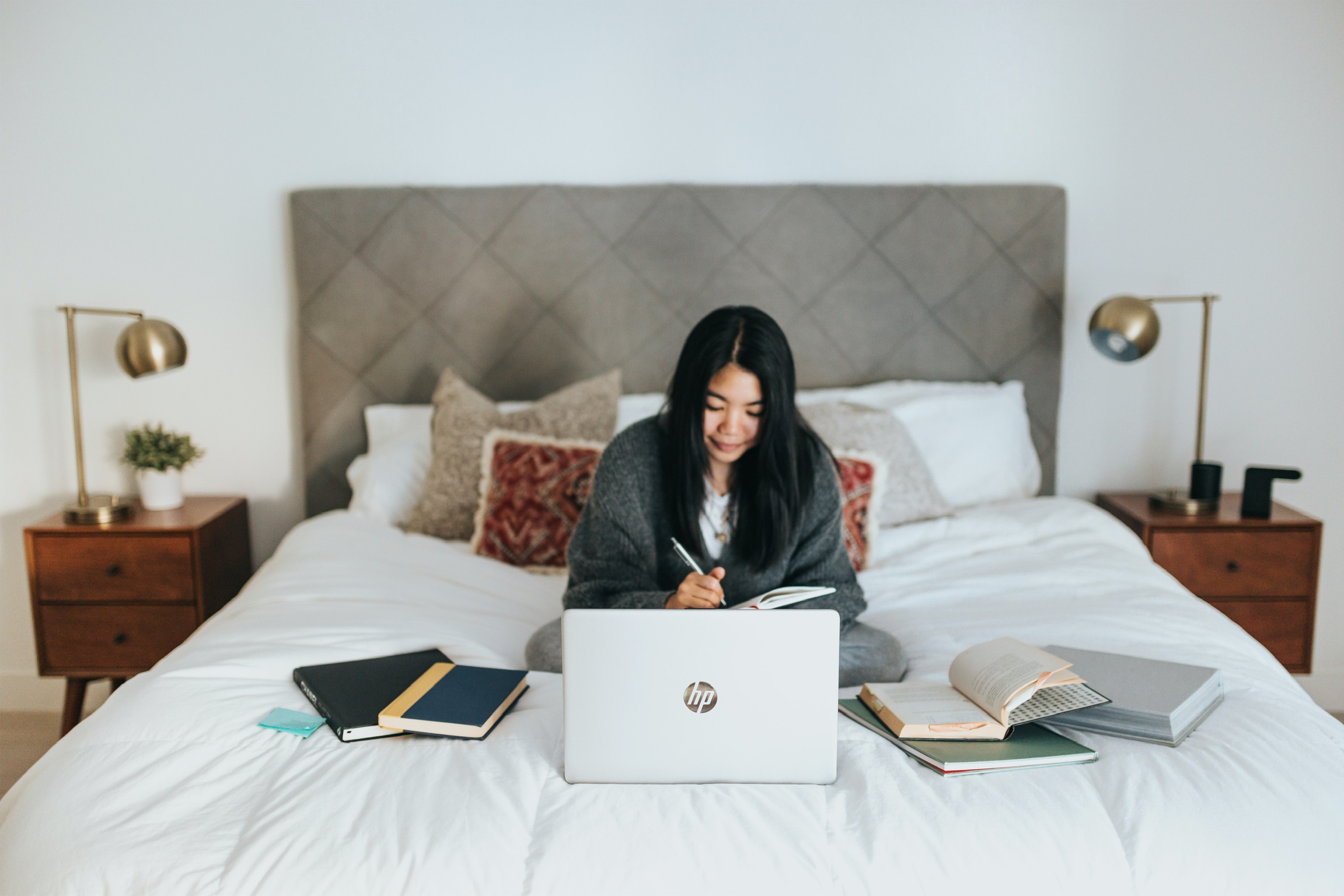 A lady working on her bedroom bend writing in a book with a laptop in front of her