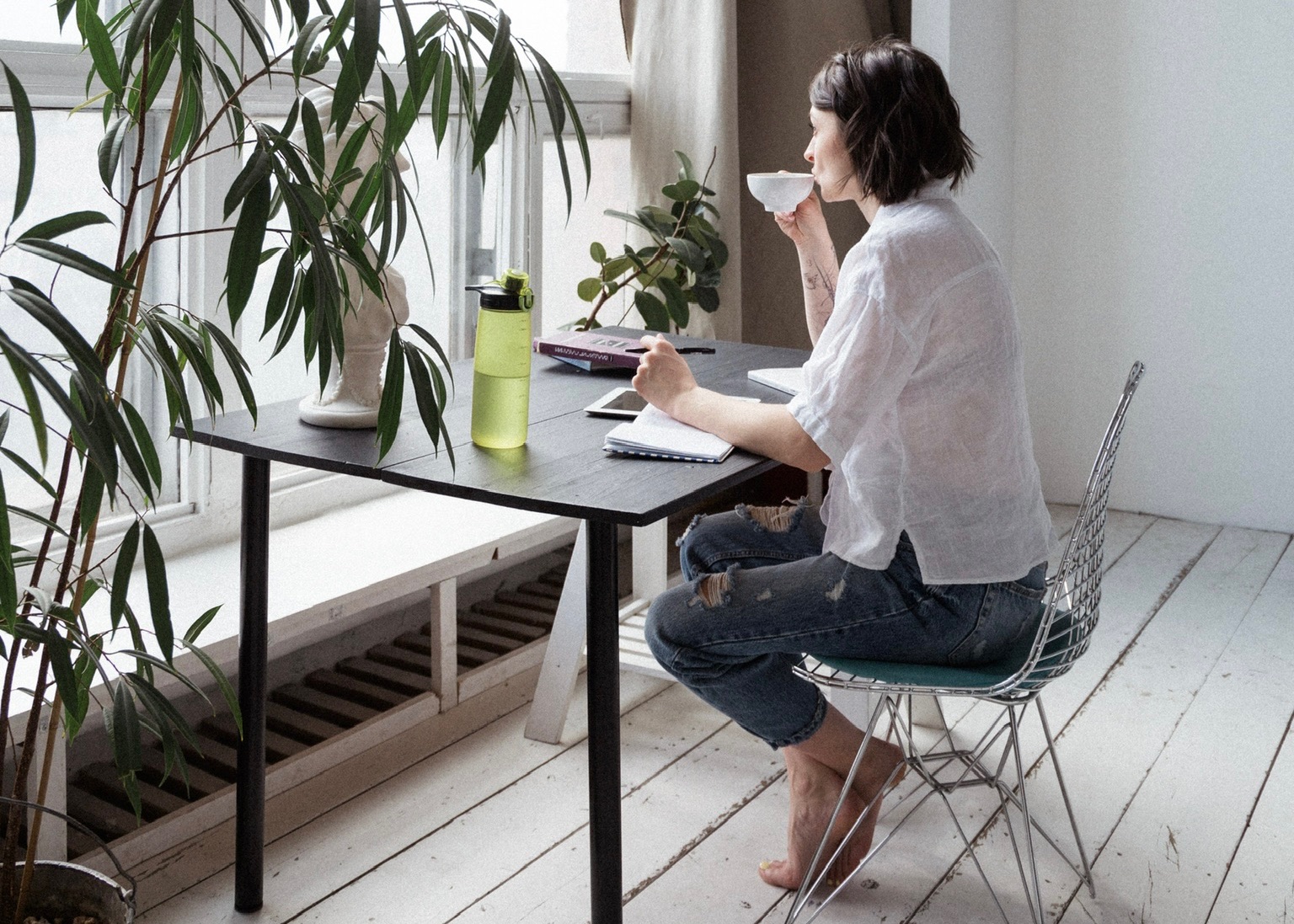 A woman at a window desk sipping a cup of coffee and holding a pen above a not book pondering looking out side the bright window