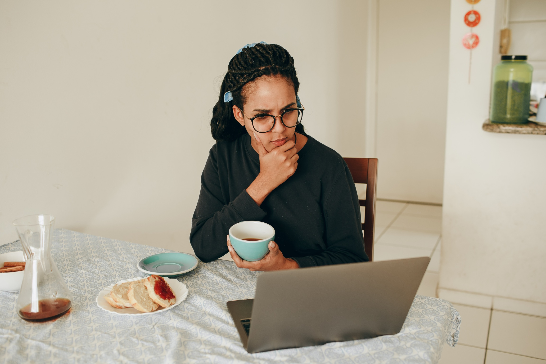 A lady sitting at a kitchen table holding her chin with one hand and a cup of tea in the other hand looking ata laptop computer